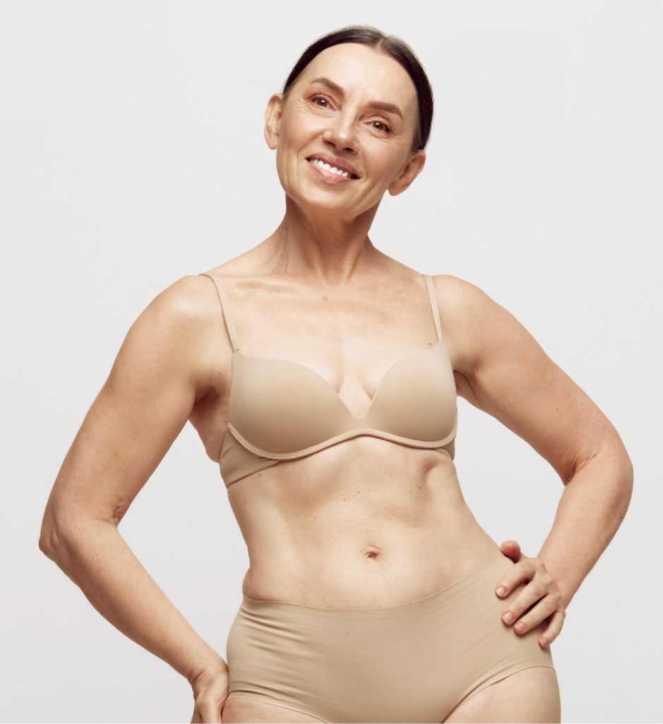 Smiling mature woman posing confidently in beige lingerie with her hands on her hips.